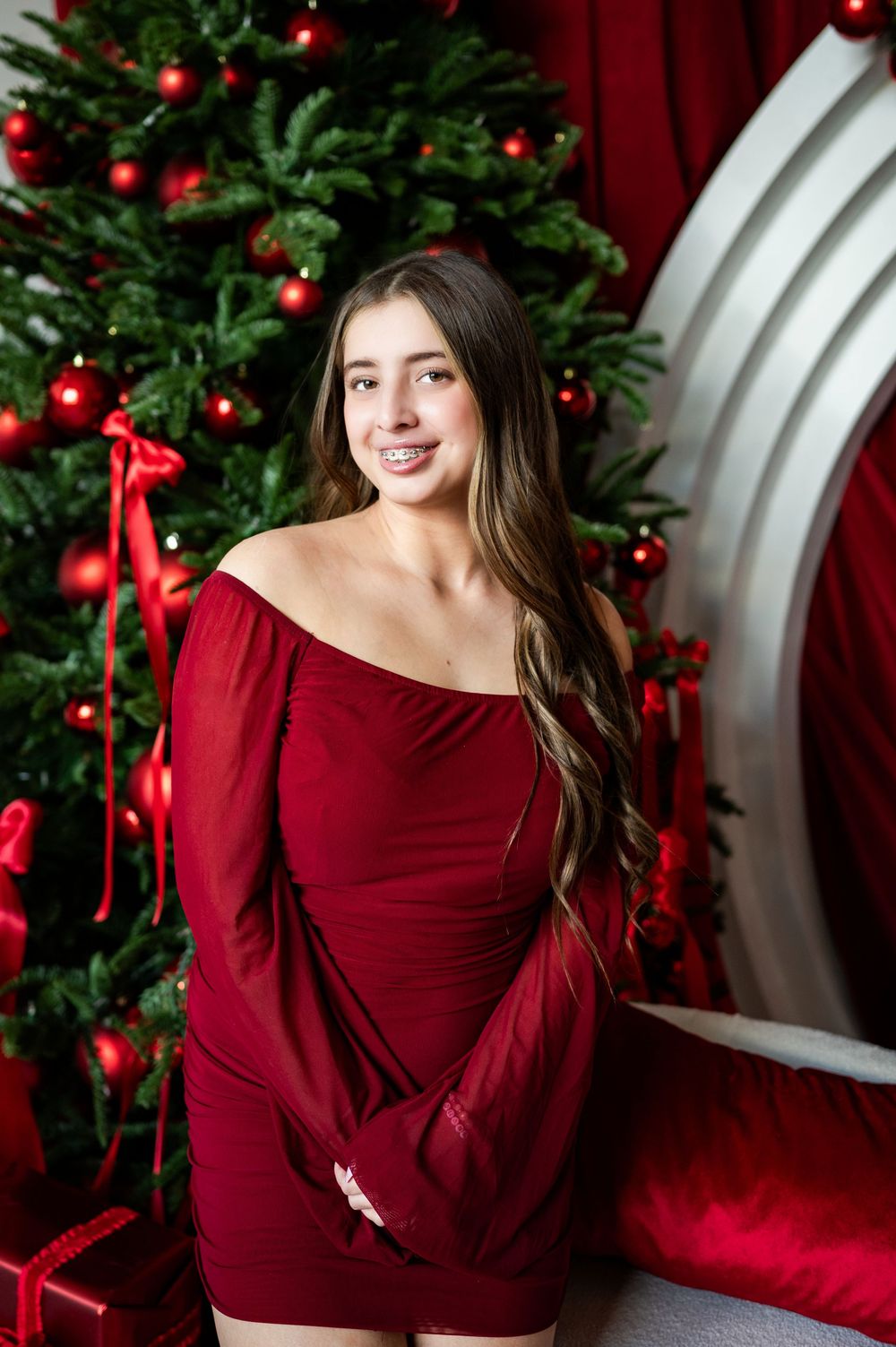 Young woman in red off-shoulder dress poses before a decorated Christmas tree with red ornaments.