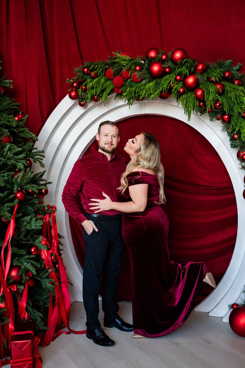 Couple in burgundy outfits posing in white circular arch with Christmas garland, red ornaments, and festive holiday decor.