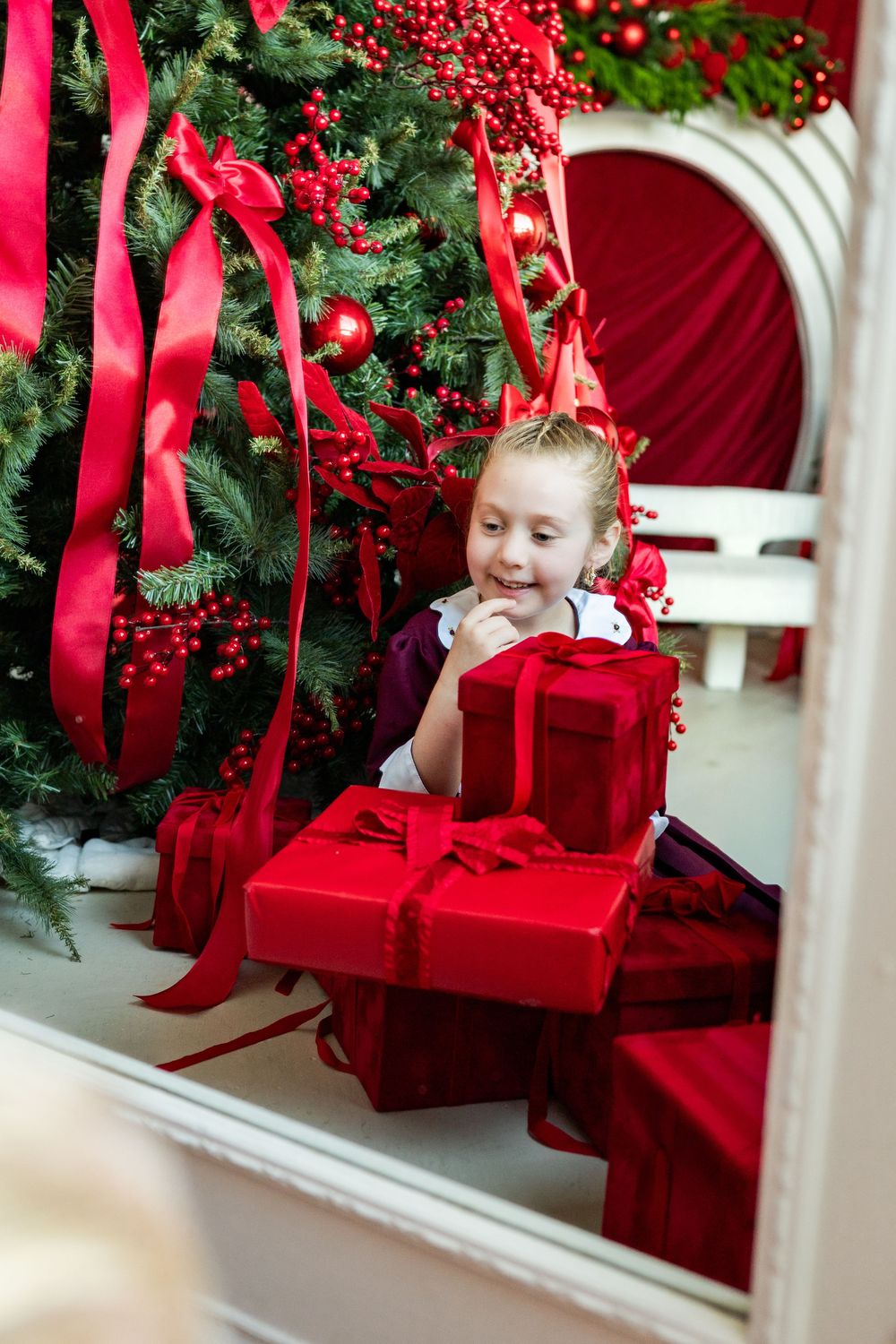 Young girl smiling with red Christmas gifts near a decorated holiday tree with red ribbons and ornaments.