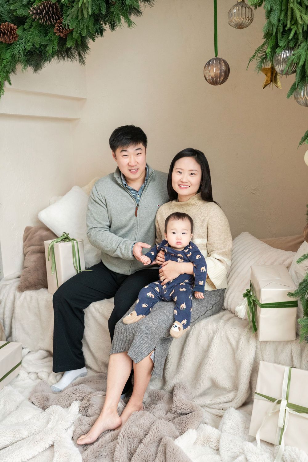 Asian family Christmas portrait with parents and baby sitting on bed surrounded by gifts and holiday greenery.