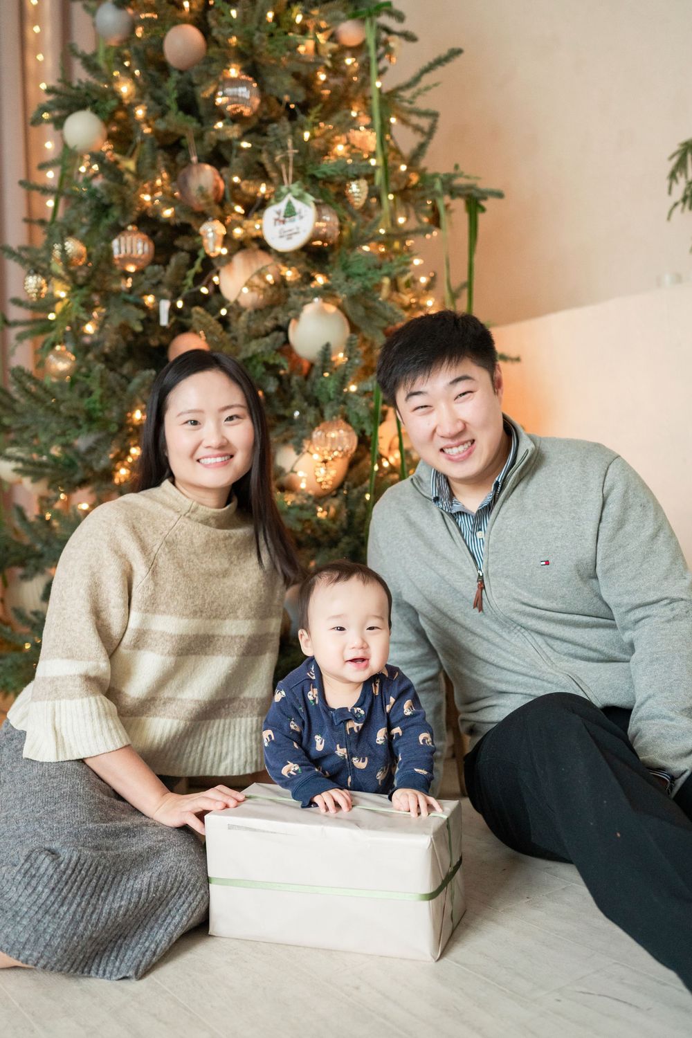 Asian family with baby smiling in front of decorated Christmas tree with gifts.