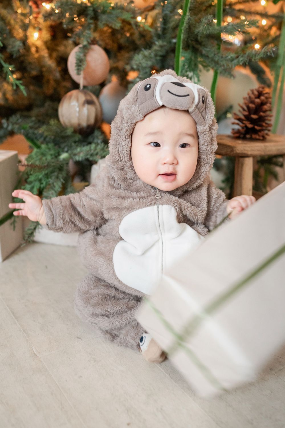 Adorable baby in a bear onesie costume sits near a decorated Christmas tree with gifts.