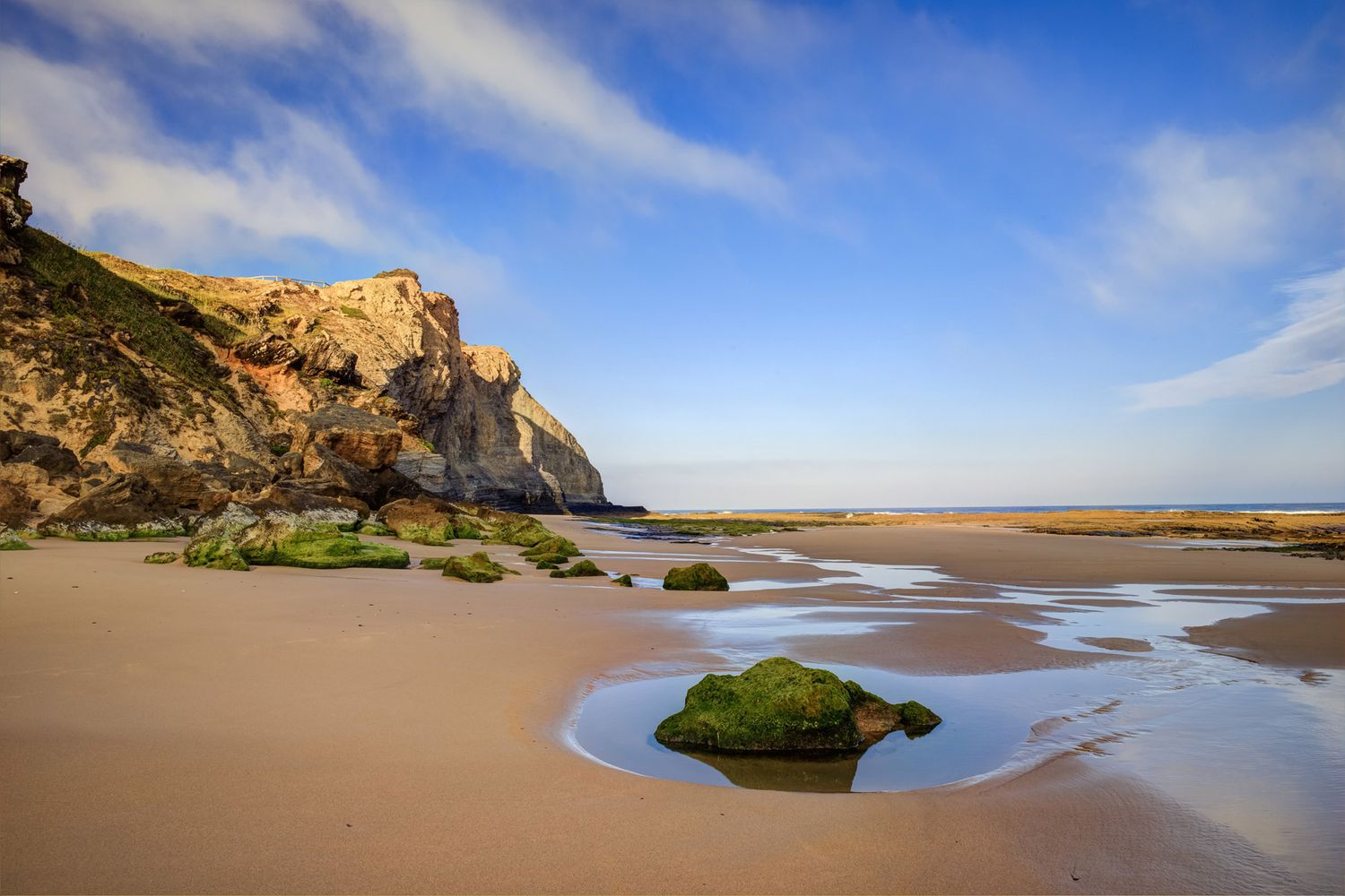 Sandy beach with moss-covered rocks, golden cliffs, and a blue sky with wispy clouds at low tide.