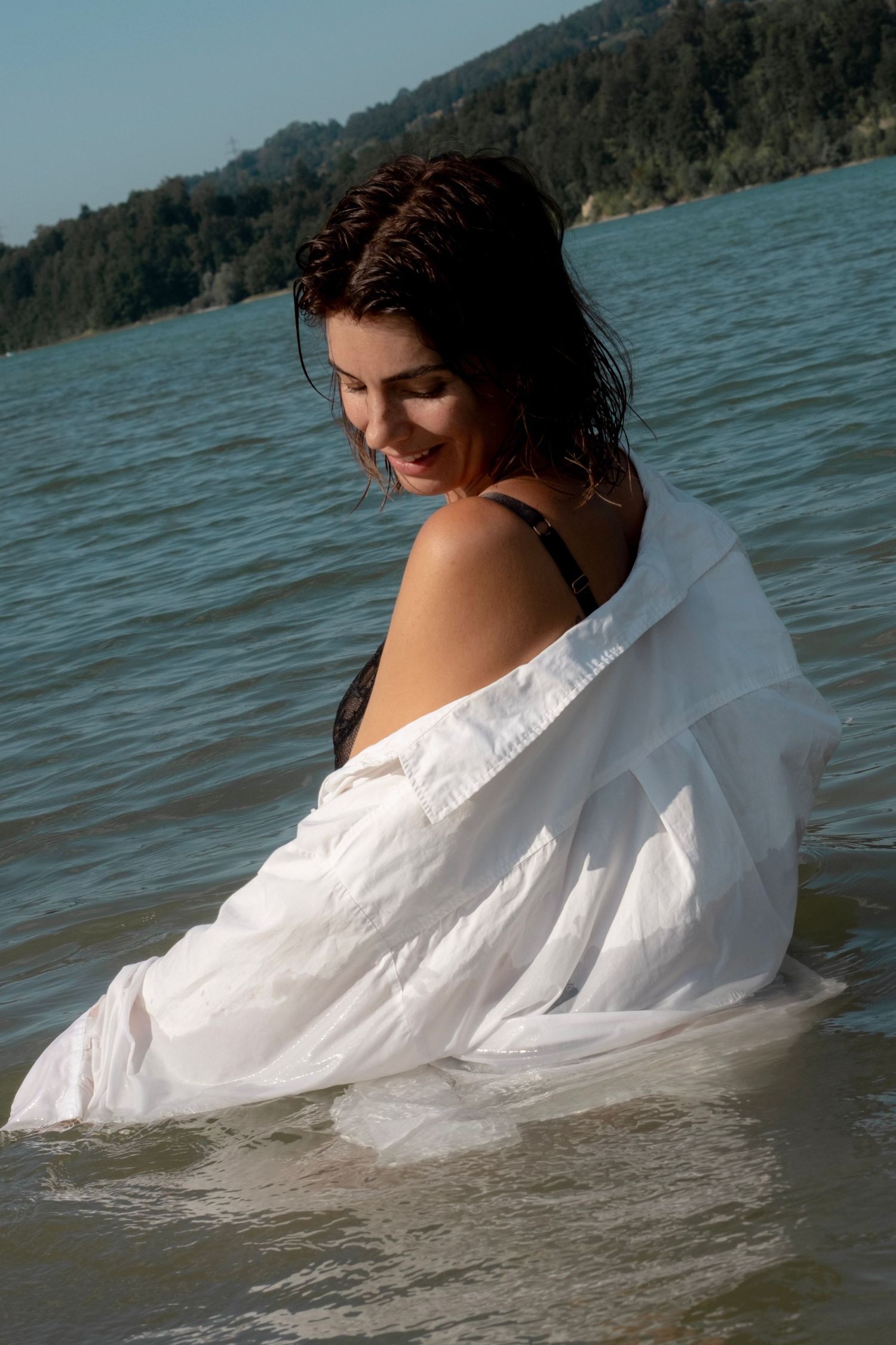 A smiling woman in a wet white shirt stands in calm ocean waters with forested hills in the background.