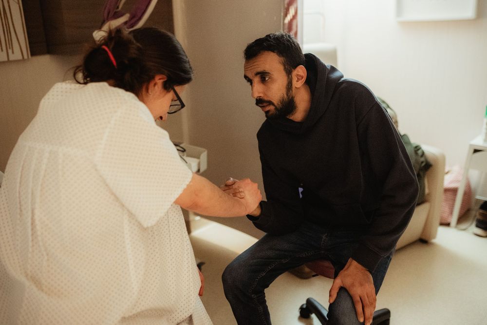 A healthcare professional examines a male patient's hand during a medical consultation in a clinic room.
