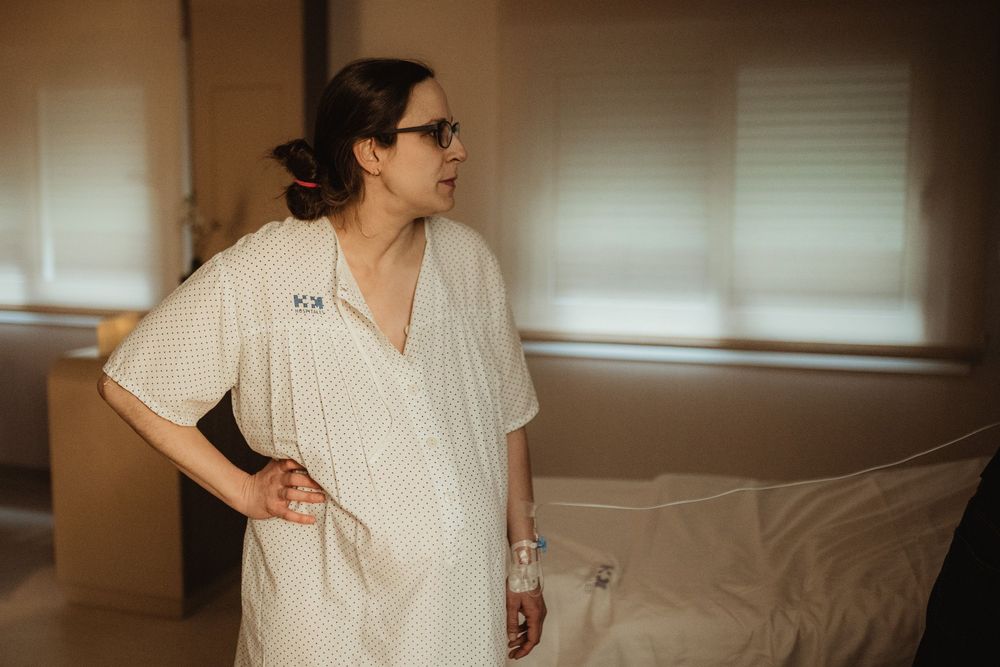 A pregnant woman in a hospital gown stands by a bed, looking toward a window in a softly lit room.