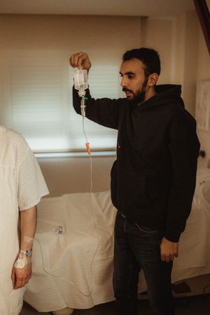 A bearded man in a black hoodie holds an IV drip bag beside a hospital patient in a medical gown.