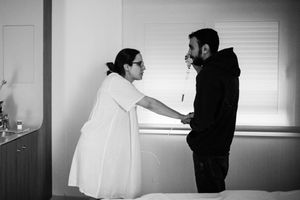 Black and white photo of a woman in white dress holding hands with a man in black near a window indoors.
