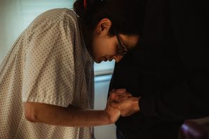 A patient in a hospital gown bends forward while a person in glasses gently holds their hands in a dimly lit room.