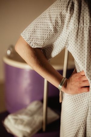 Patient in hospital gown with ID wristband standing beside medical equipment in a clinical room.