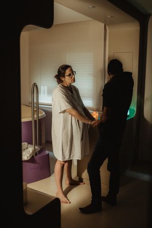 A pregnant woman in a hospital gown holds hands with her partner in a dimly lit birthing suite.