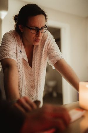 A focused woman with glasses in a white blouse performs a massage therapy session in a warmly lit room.