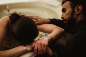 A man tenderly supports a woman leaning over a birthing tub during labor, holding her hand gently.