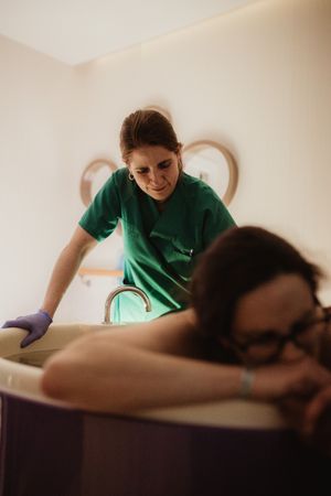 A midwife in green scrubs assists a woman during a water birth in a birthing tub.