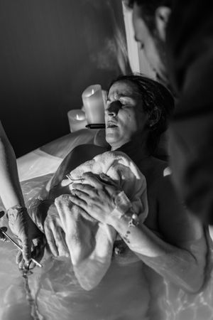 Black and white photo of a woman holding her newborn baby during a water birth, with candles glowing softly in the background.