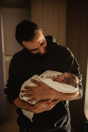 A father tenderly holds his newborn baby wrapped in a white blanket in a warmly lit room.