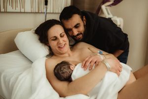 New parents smile joyfully in hospital bed, mother holding newborn baby skin-to-skin after birth.