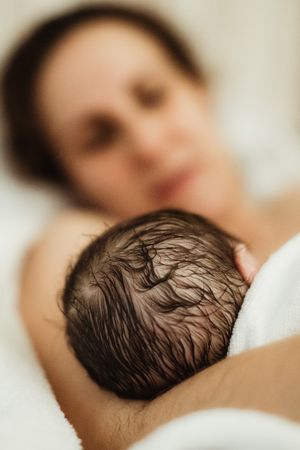 A mother tenderly holds her newborn baby with dark wet hair wrapped in a white blanket after birth.