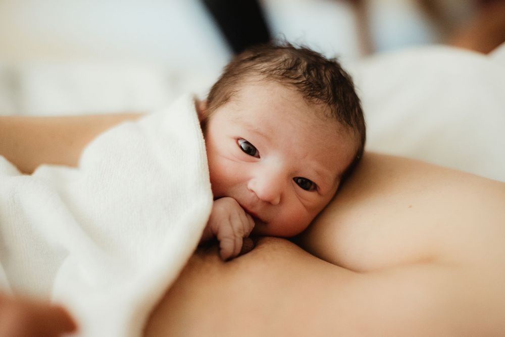 Newborn baby breastfeeding, wrapped in white blanket, looking up with dark eyes in a tender moment.