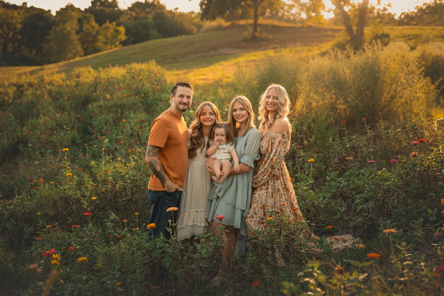Family of five posing in a golden sunlit meadow with wildflowers, smiling warmly during an outdoor photo session.