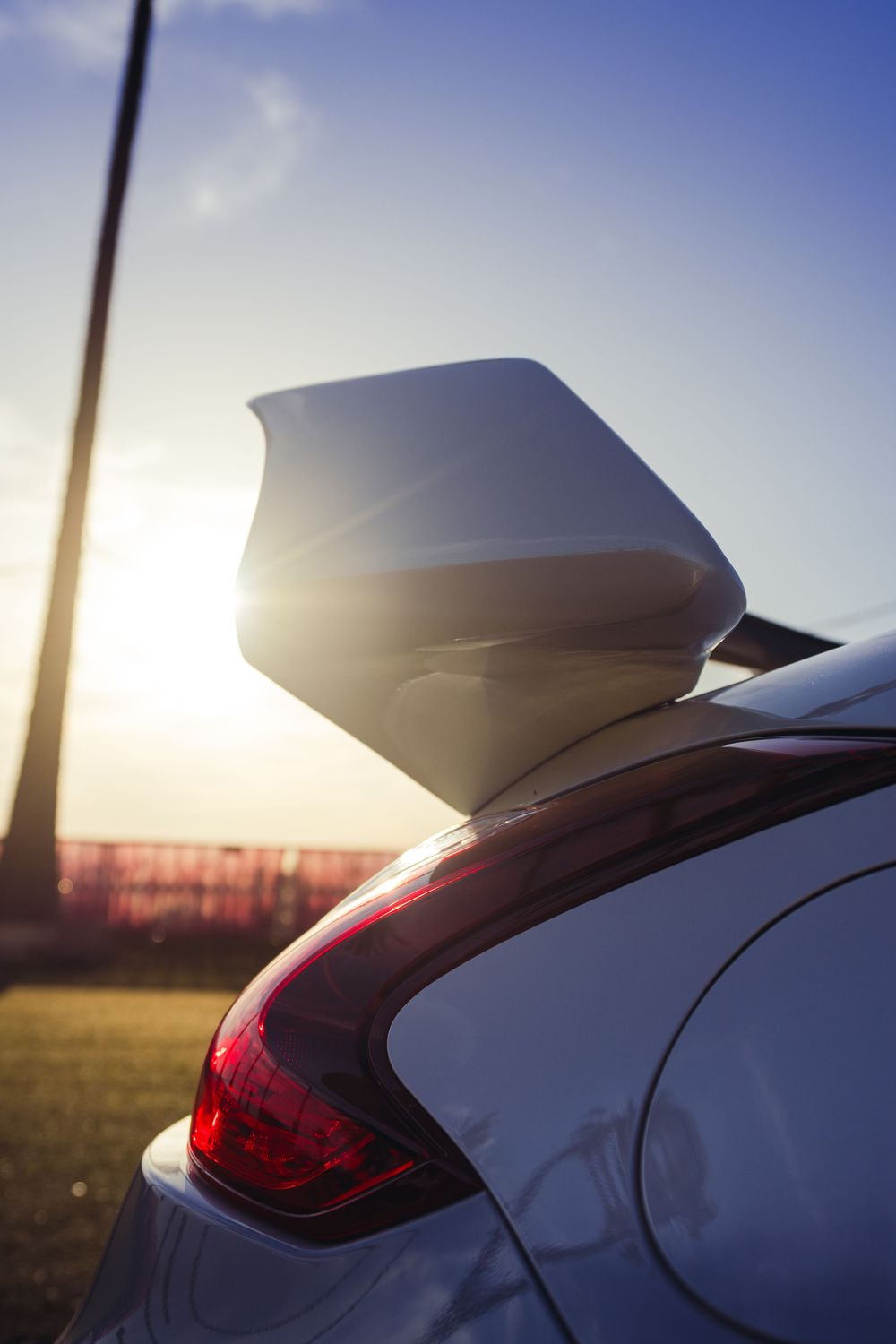 Close-up of white sports car rear spoiler and taillight glowing against a vibrant sunset sky.