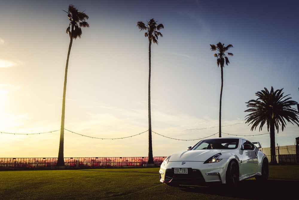 White Nissan 370Z Nismo parked on grass near palm trees at sunset by the ocean.