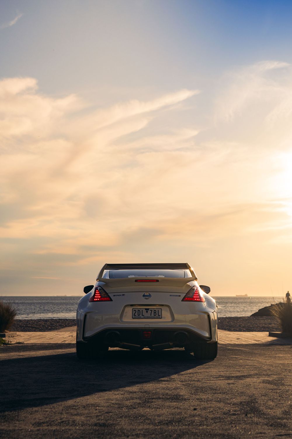 Rear view of a white Nissan 370Z Nismo parked by the ocean at golden hour sunset.