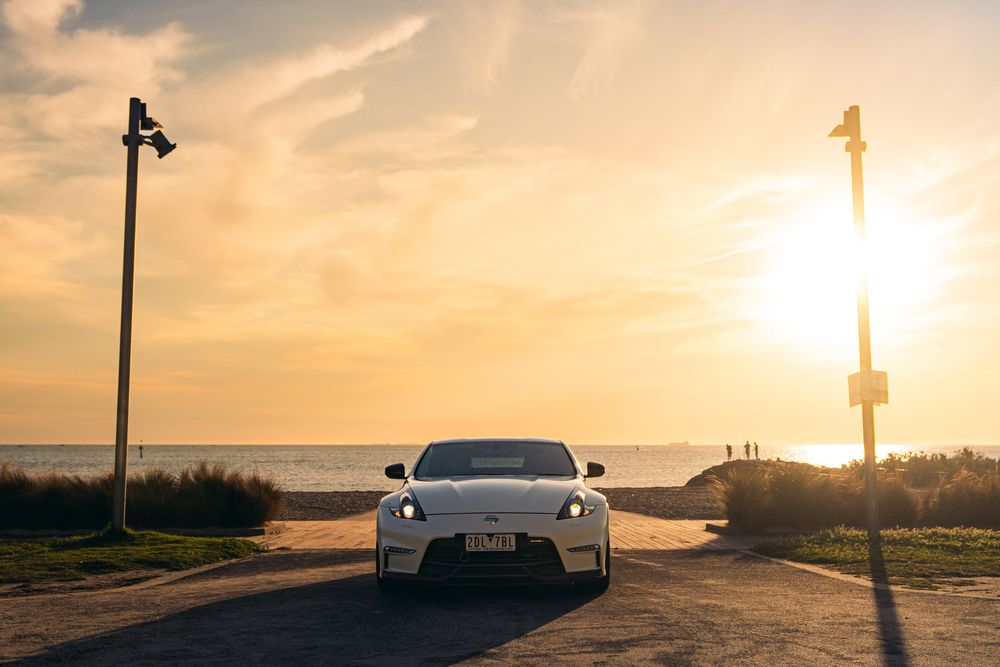 White Nissan 370Z parked near the ocean at golden sunset with lamp posts framing the scene.