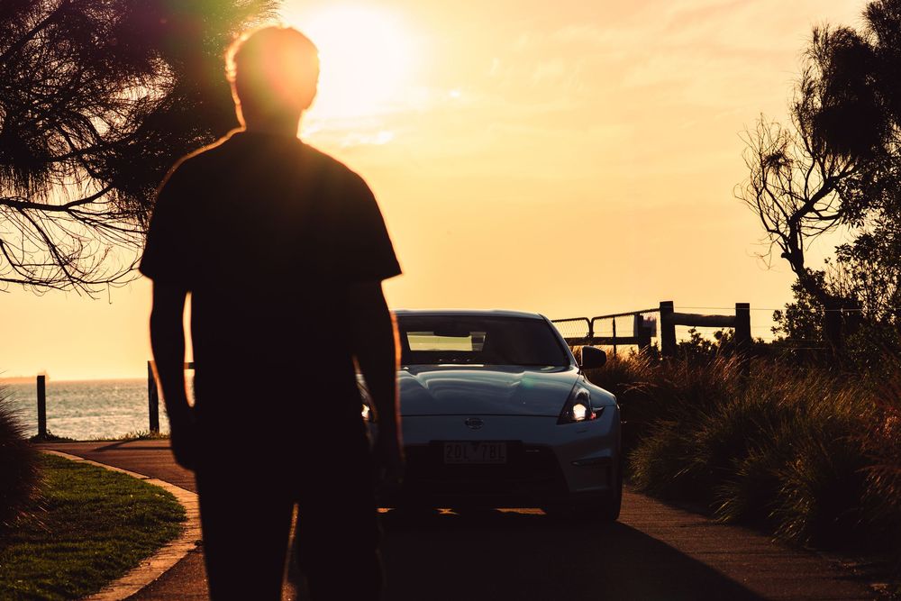 Man standing in front of white Nissan 370Z sports car at stunning ocean sunset.