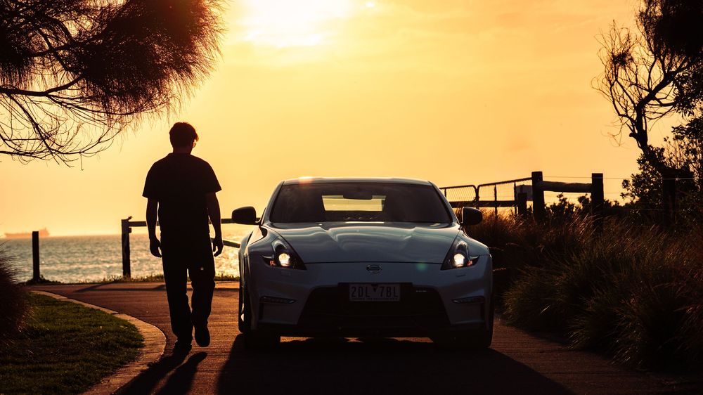 Silhouette of man walking beside white Nissan 370Z on coastal road at sunset.