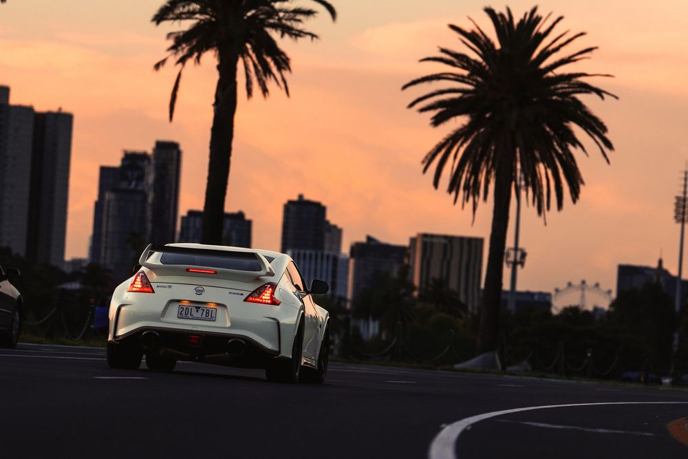 White Nissan 370Z Nismo driving past palm trees with city skyline at dusk.