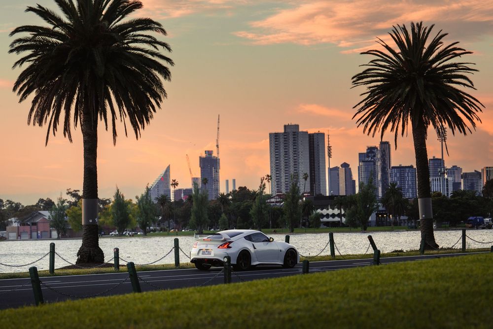 White Nissan 370Z parked between palm trees with Melbourne city skyline at sunset.