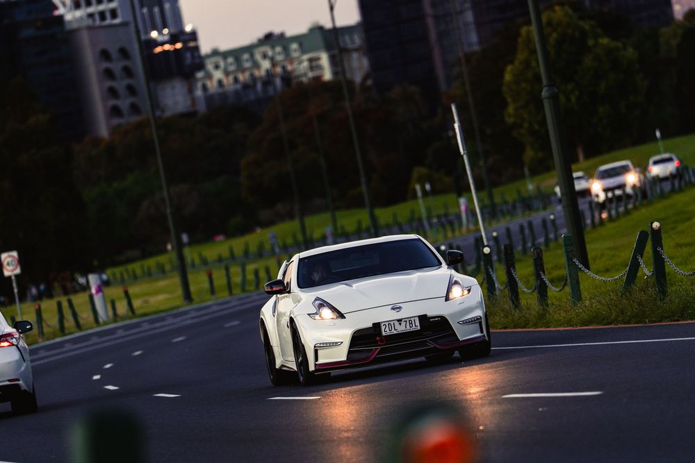 White Nissan 370Z Nismo driving on urban road with city buildings in background at dusk.