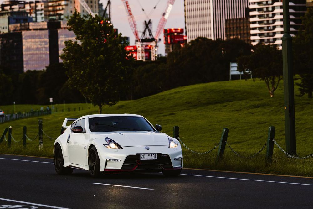 White Nissan 370Z Nismo driving on urban road with city skyline and cranes in background.
