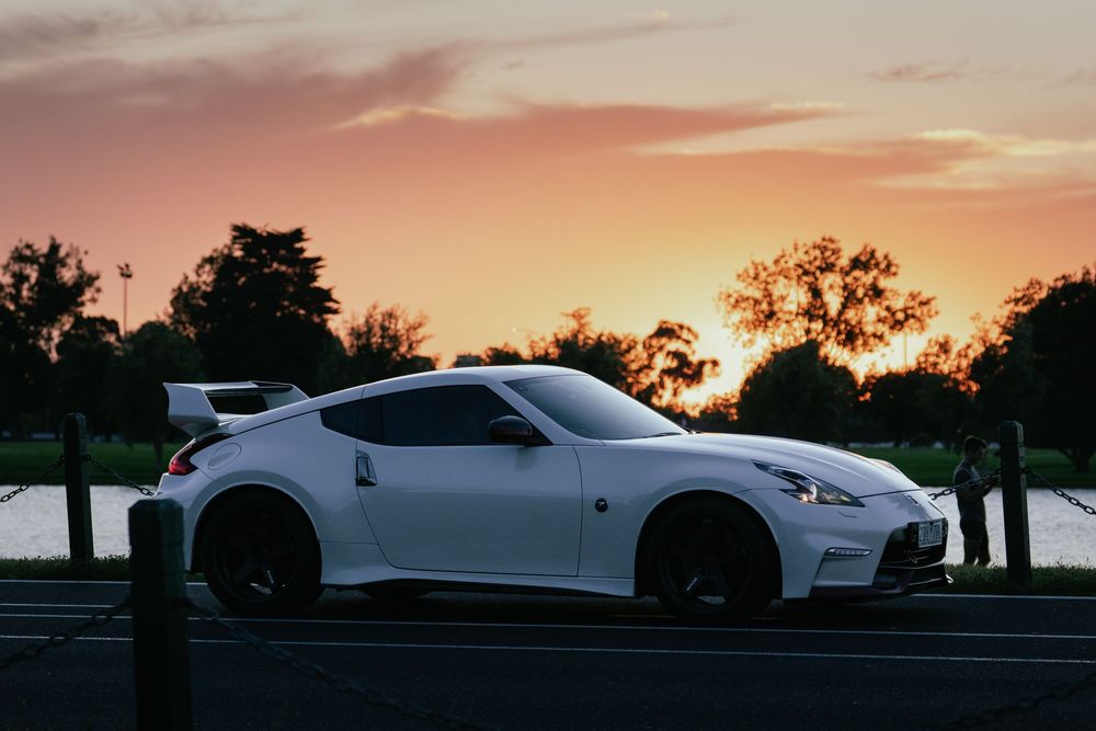 White Nissan 370Z Nismo parked beside lake with vibrant orange and pink sunset sky.