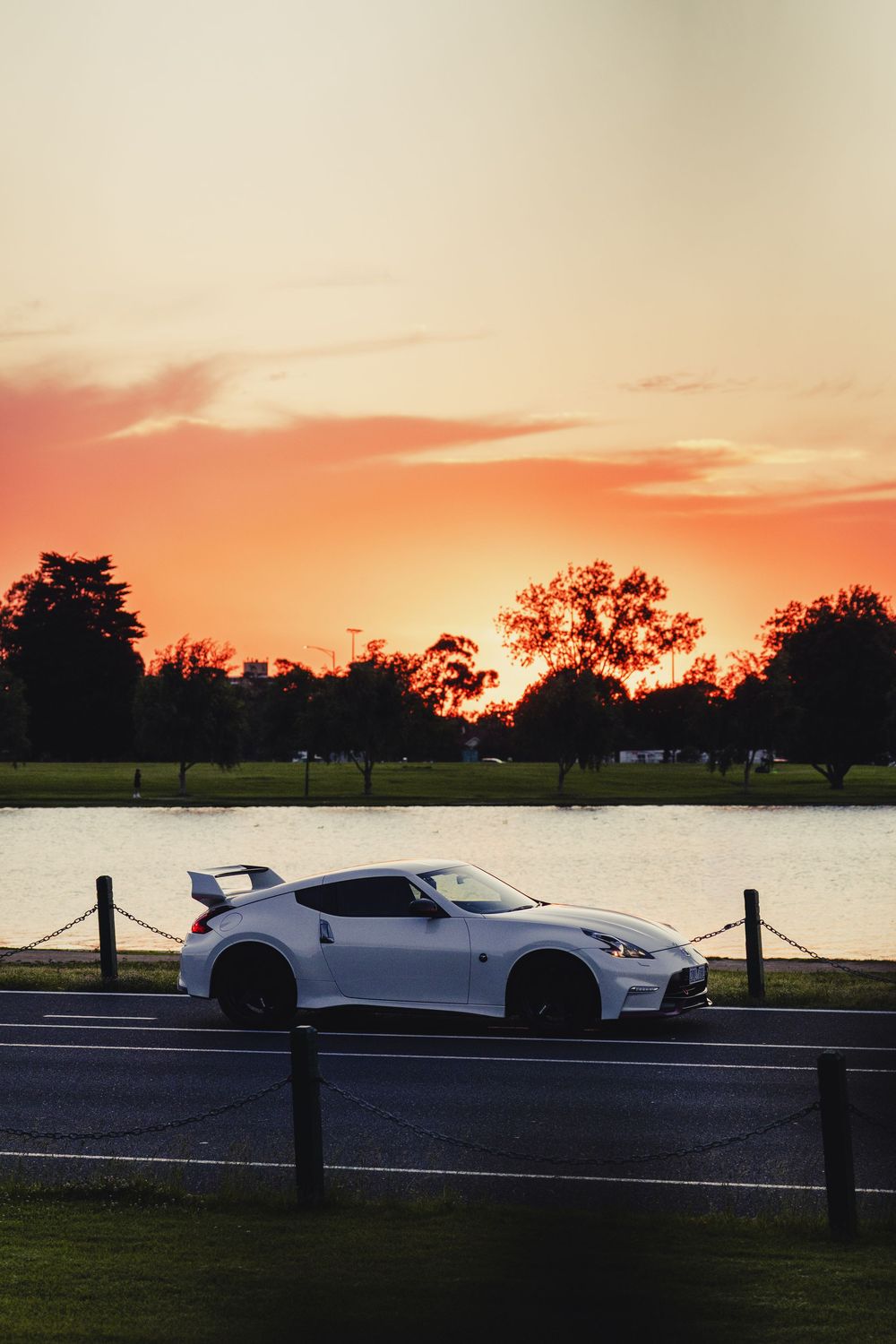 White Nissan 370Z Nismo parked roadside with stunning orange sunset reflecting over calm lake.