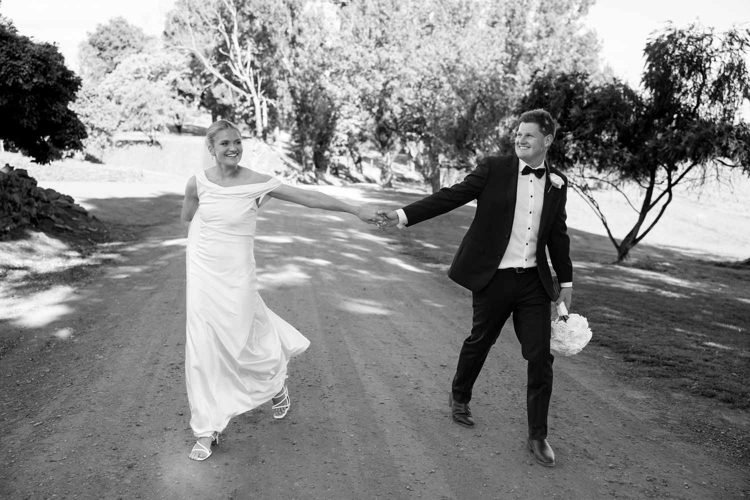 Black and white photo of a bride and groom joyfully holding hands while walking on a sunlit dirt path.