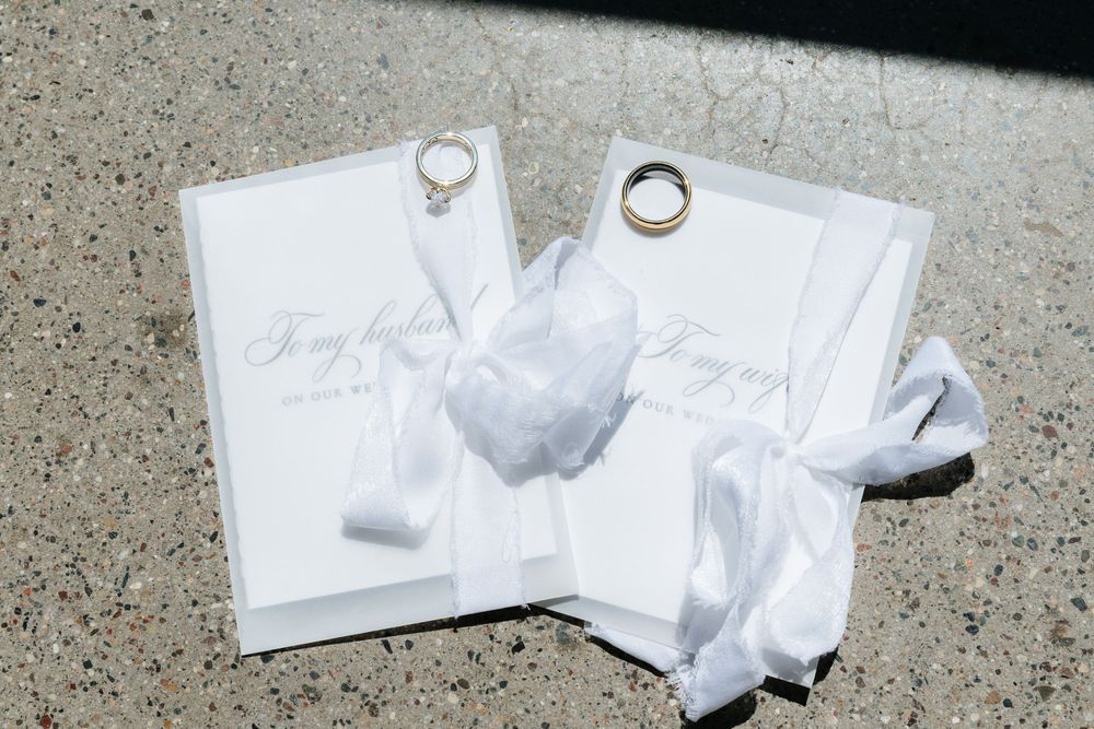 Two white wedding vow books with satin ribbons and gold wedding bands displayed on a textured surface in natural light.