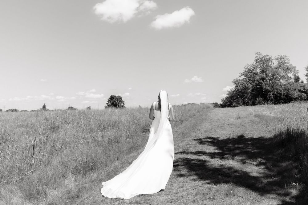 Black and white photo of a bride in a flowing white gown and veil standing in a scenic field on her wedding day.