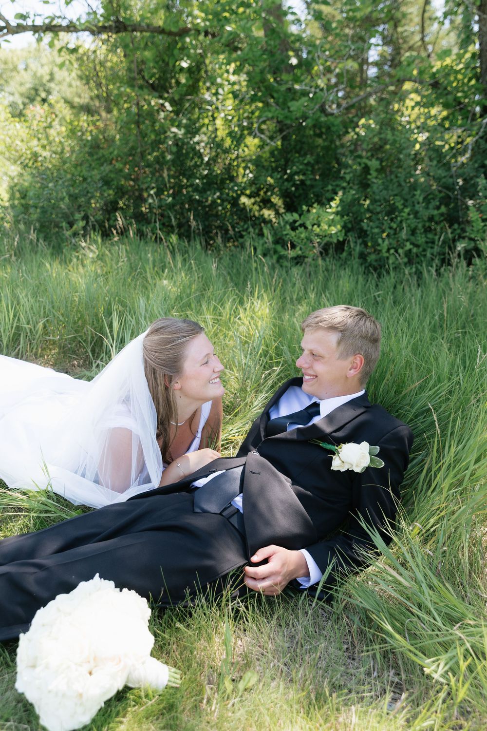 Bride and groom lying in lush green grass, smiling at each other on their wedding day.