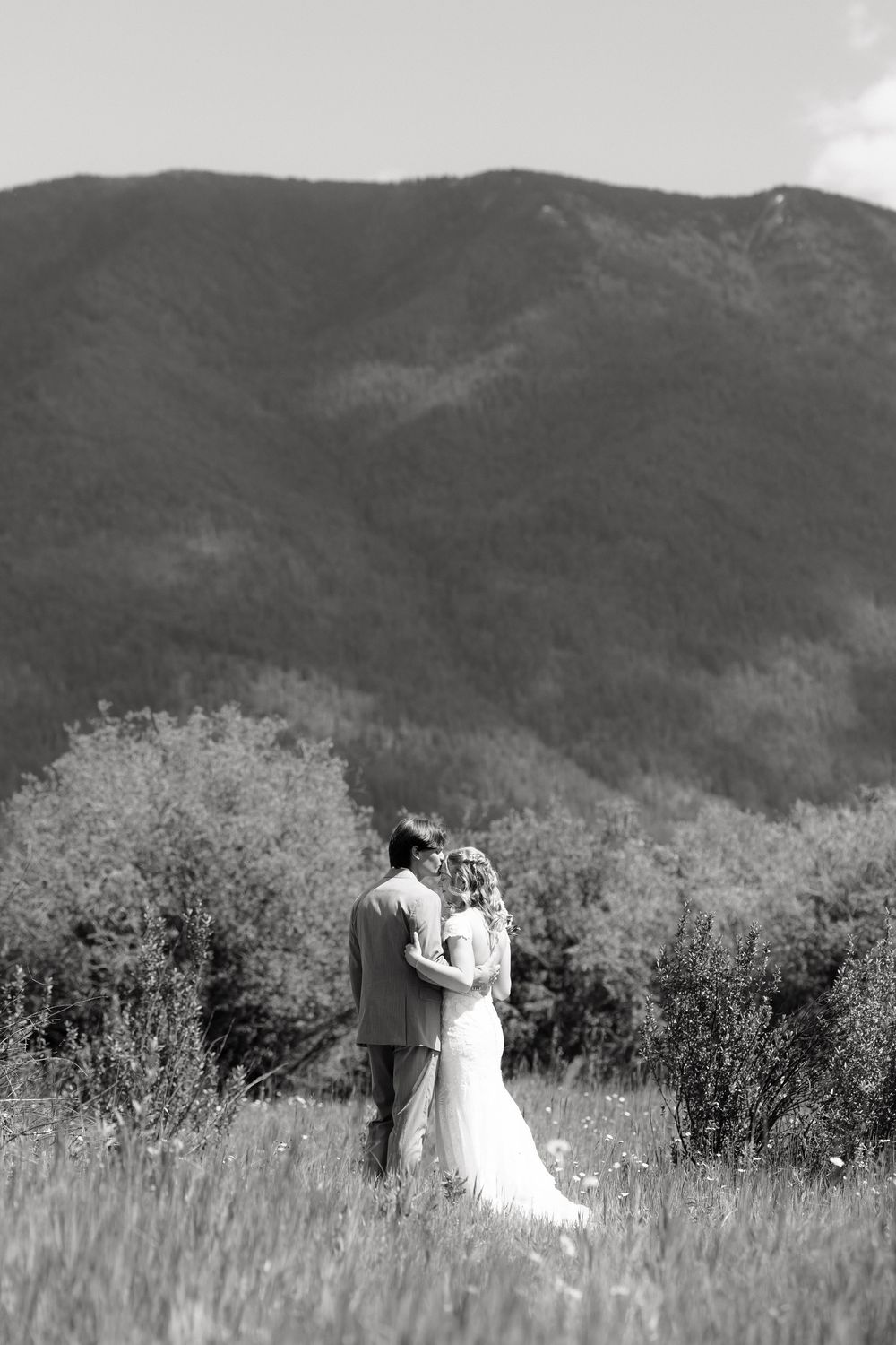 Black and white photo of a bride and groom embracing in a meadow with a lush mountain backdrop.
