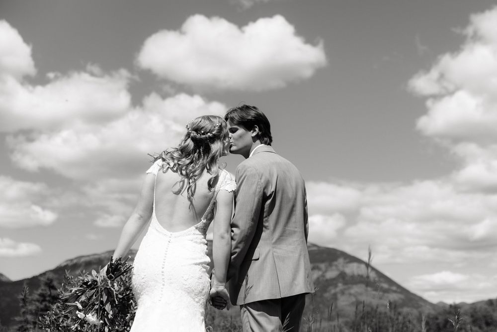 Black and white photo of a bride and groom kissing with mountain landscape and cloudy sky backdrop.
