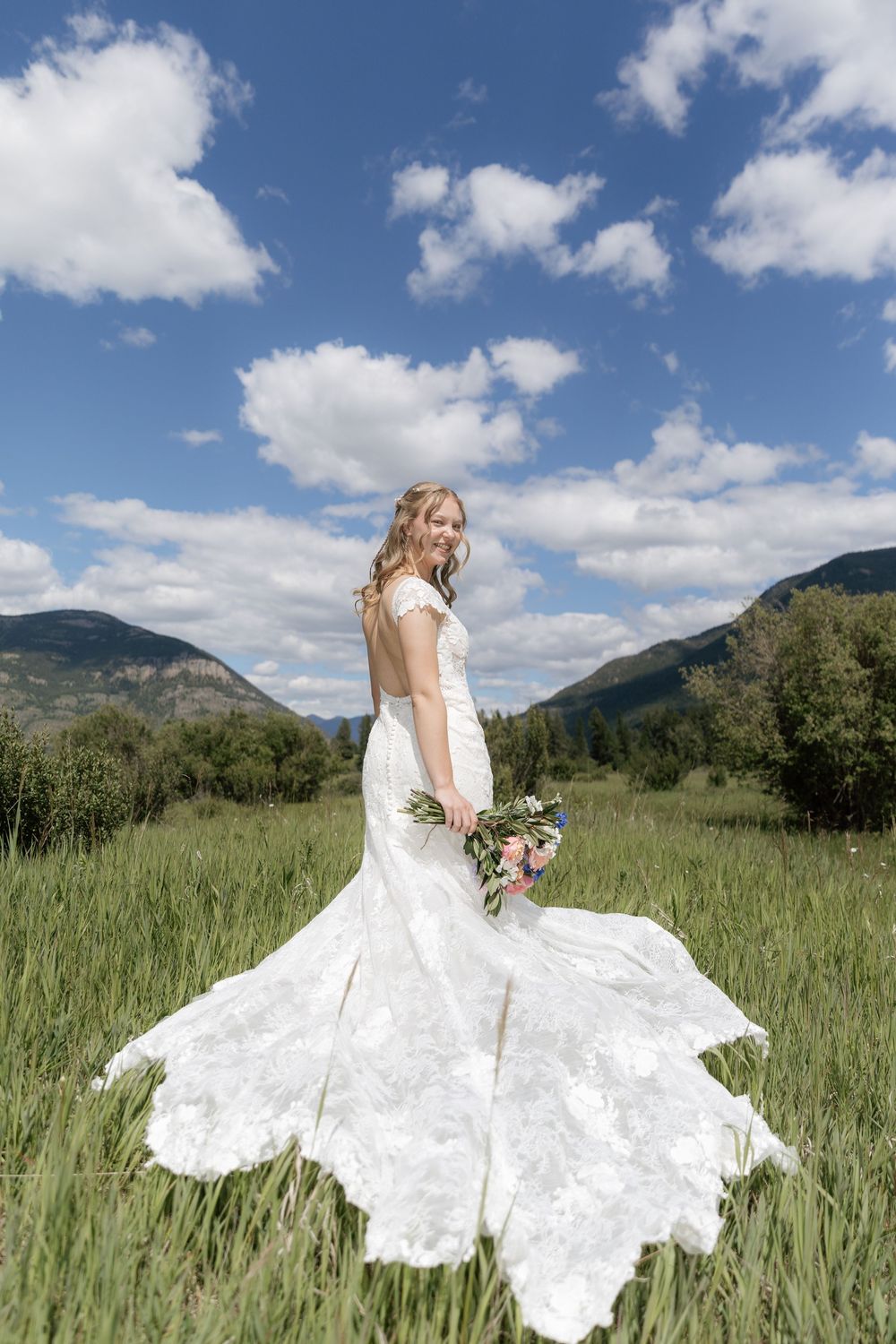 Bride in white lace gown with train holds bouquet in green mountain meadow under blue sky.