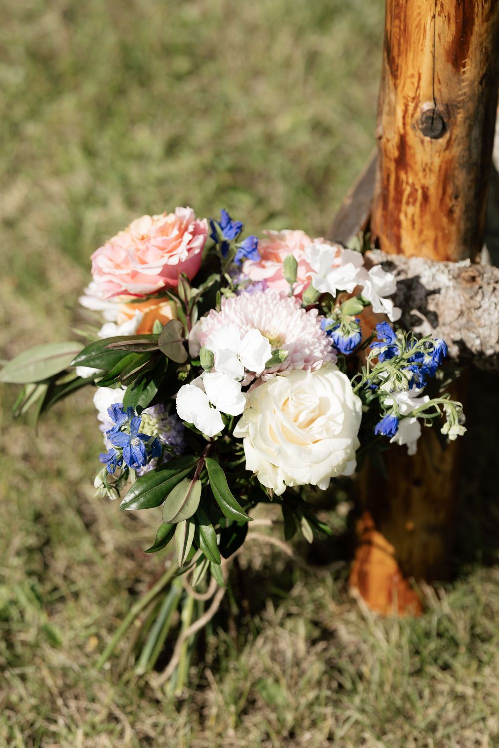 Rustic wedding bouquet with white and pink roses, blue flowers, and greenery leaning against a wooden post outdoors.