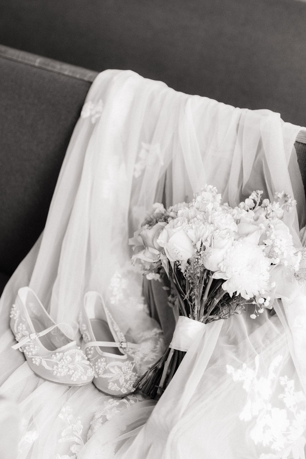 Black and white photo of bridal accessories including lace heels, floral bouquet, and flowing veil on a sofa.