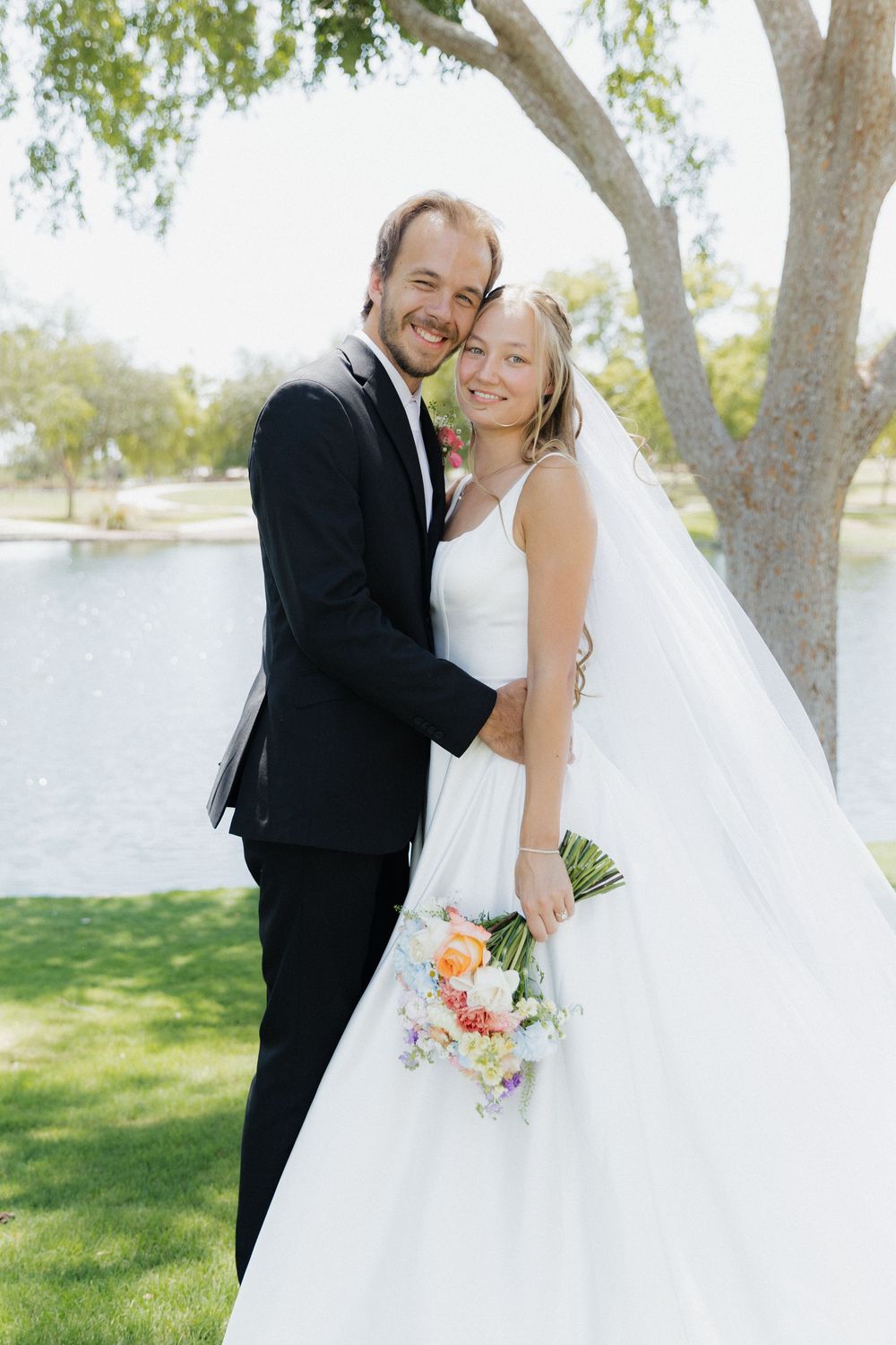 Smiling bride and groom embracing outdoors near a pond, bride holding a colorful bouquet in a white gown.