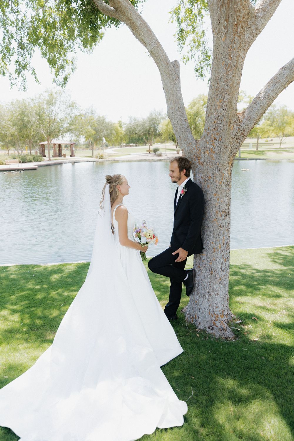 Bride in white gown and groom in black suit share a romantic moment by a tree near a serene pond.