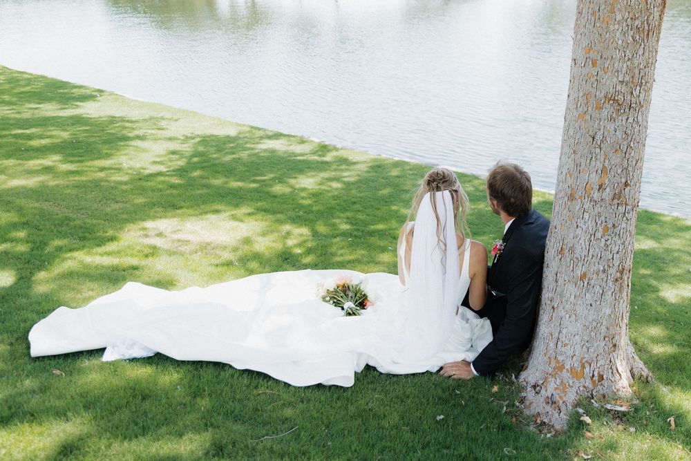 Bride and groom sitting together by a tree near a serene lake, with wedding dress train spread on green grass.