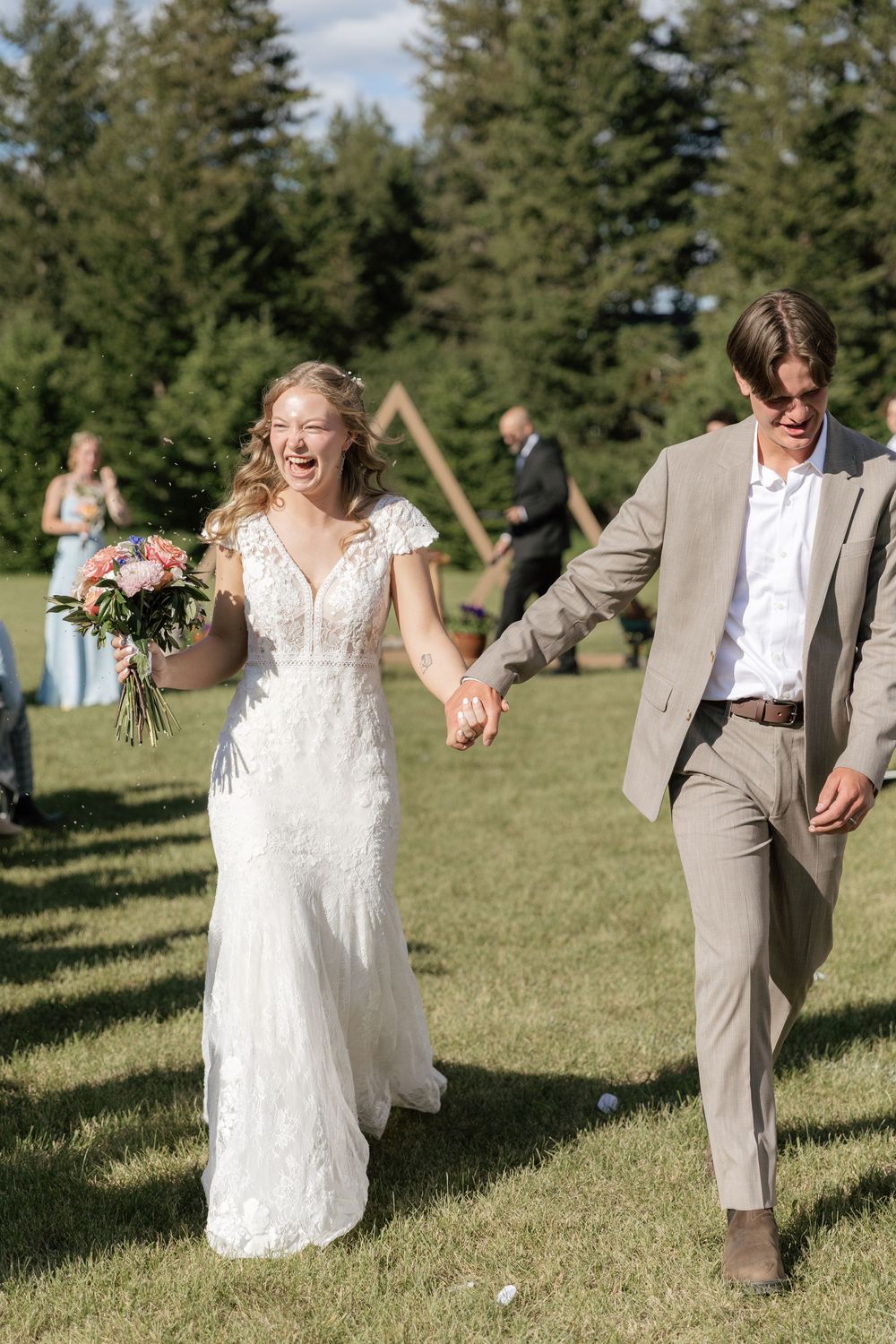 Joyful newlyweds walk hand-in-hand through outdoor wedding venue, bride in lace gown holding bouquet, groom in tan suit.