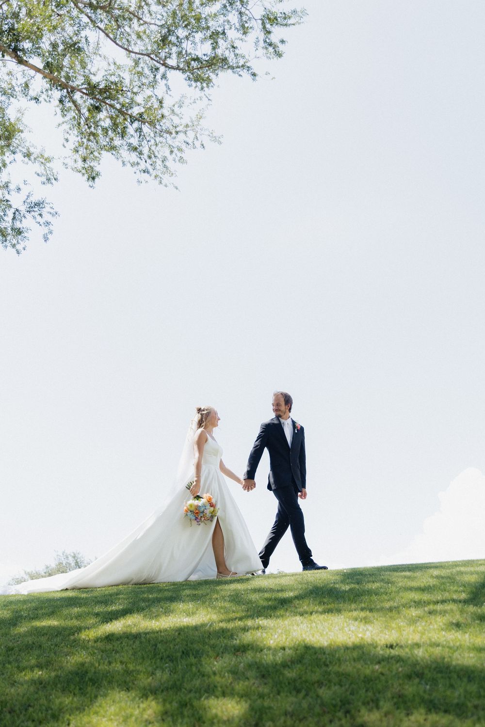 Bride and groom holding hands walking on green grass hill under tree on sunny wedding day.